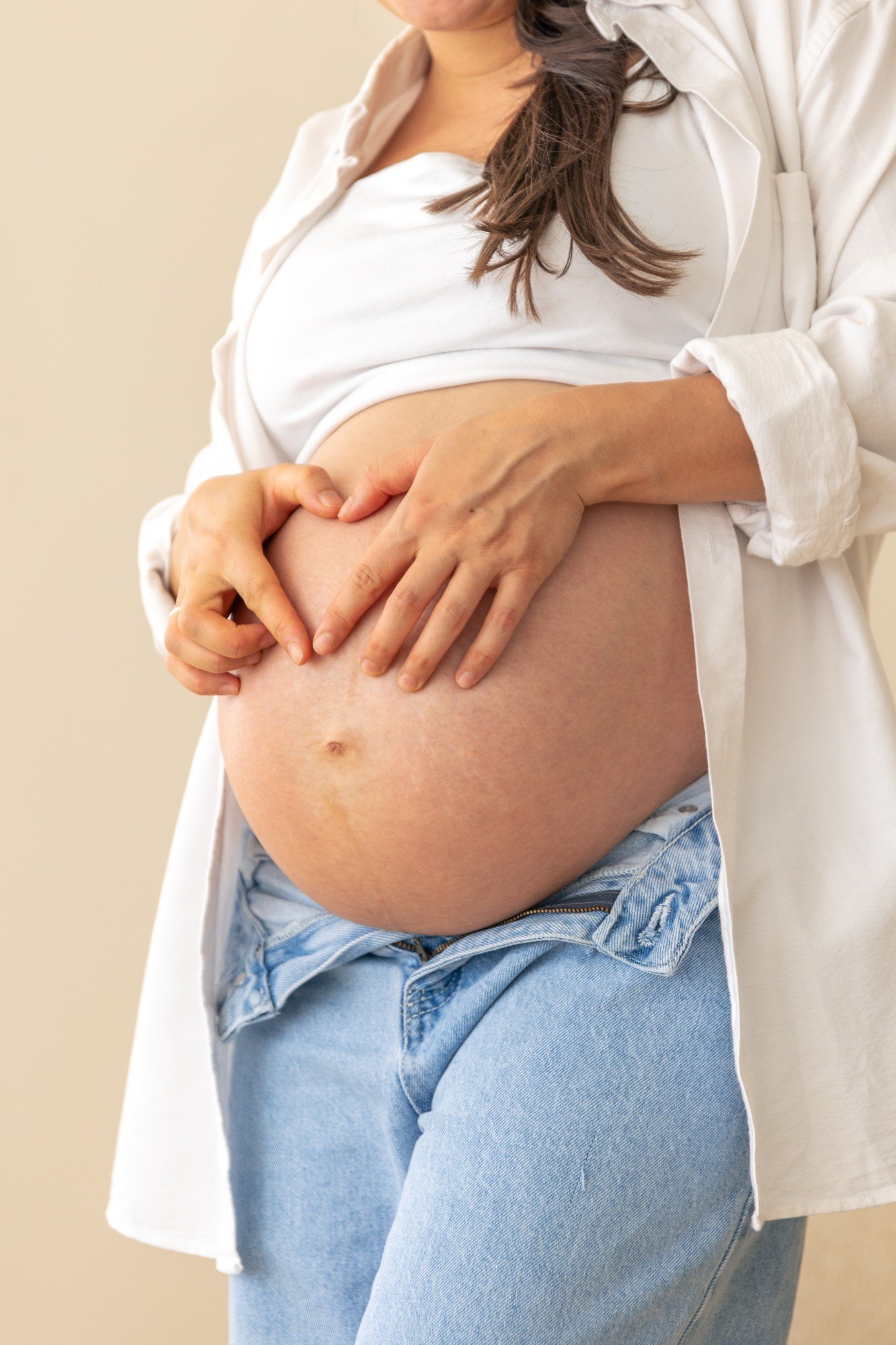 Gestante de camisa branca e calça jeans, com as mãos formando um coração sobre a barriga